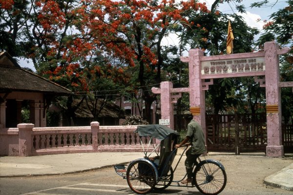 "Vietnam Hué. Rue de Hué, rive droite". Un cyclo-pousse passe devant une porte monumentale de couleur rose